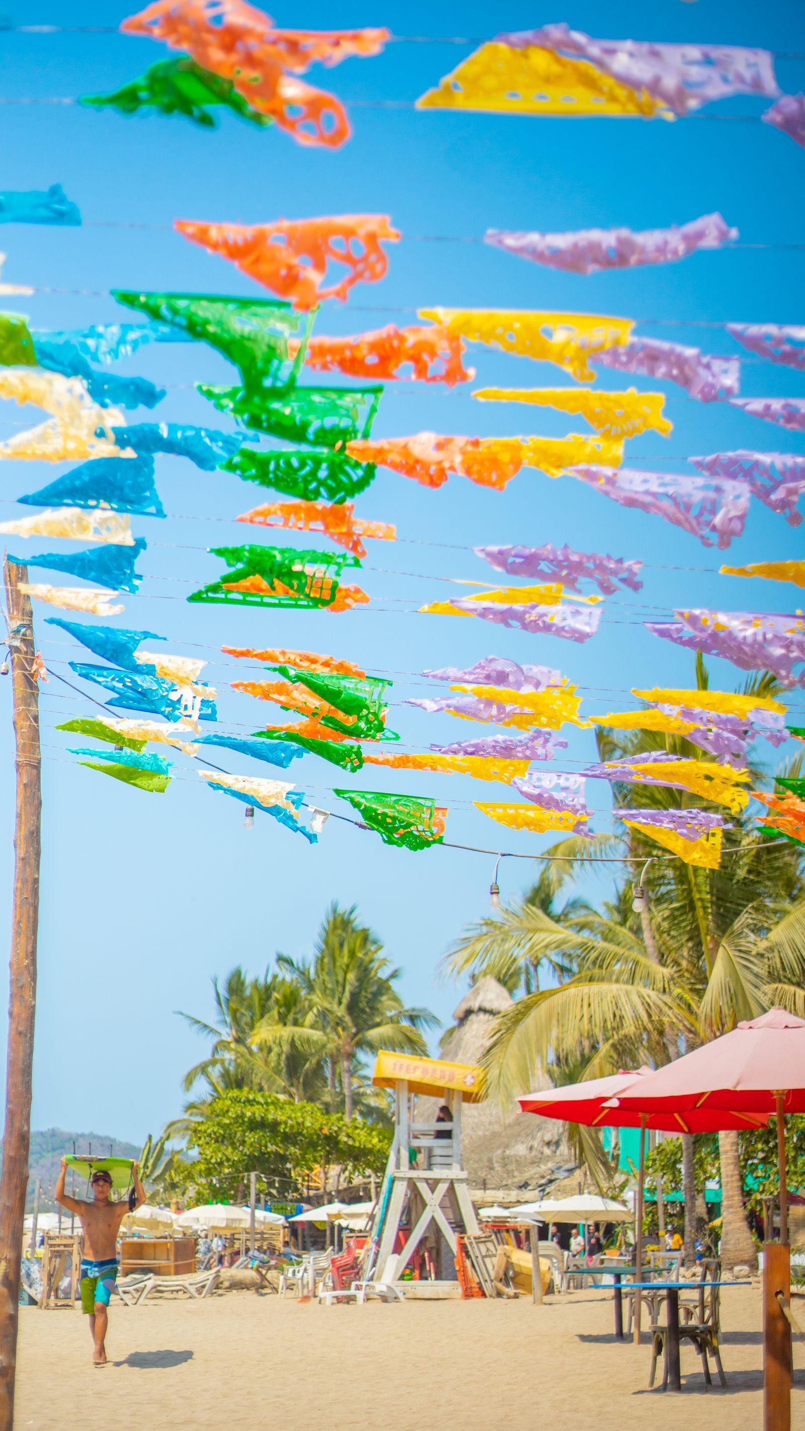 Papel picado de colores sobre la playa de Sayulita, con una torre de salvavidas y un surfista al fondo