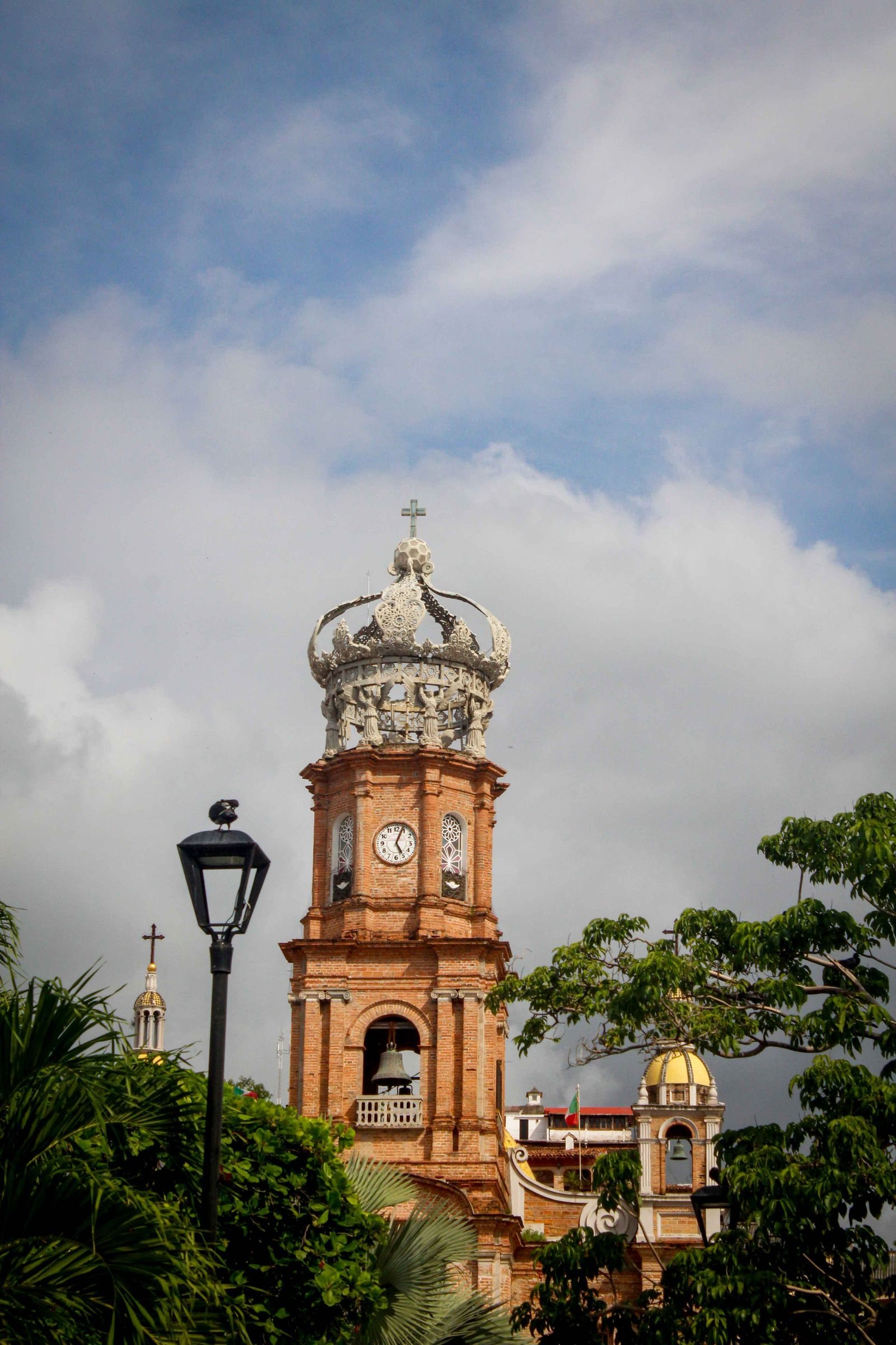 Torre de la Parroquia de Guadalupe con su corona, campanario y reloj entre árboles y un farol
