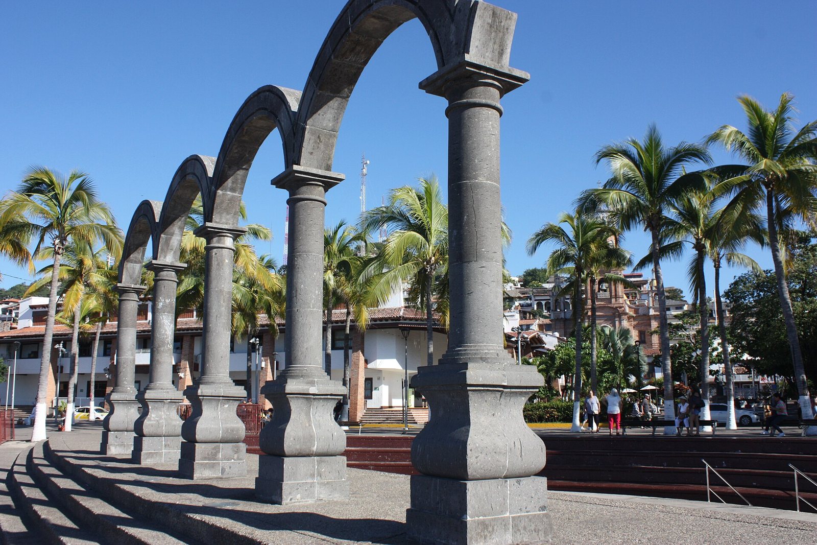 Cuatro arcos de cantera gris del anfiteatro Los Arcos en el Malecón, con palmeras y la torre de la parroquia al fondo