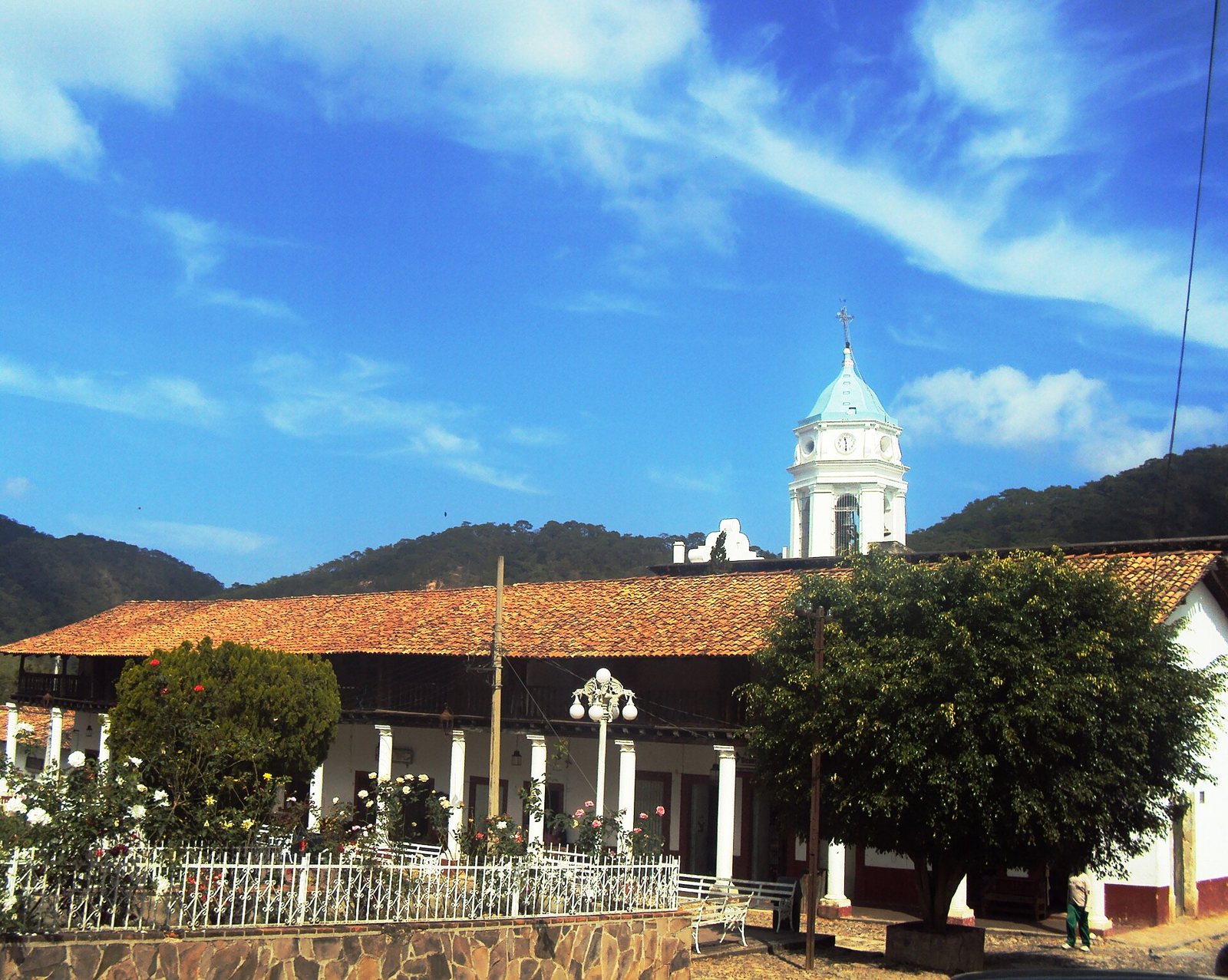 Torre blanca del templo de San Sebastián y portales con techo de teja, con cerros boscosos al fondo