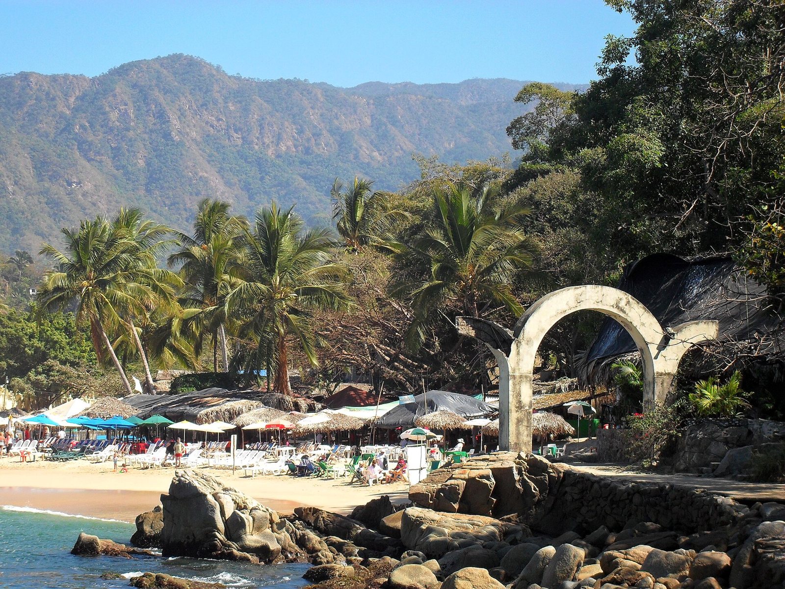Palapas y camastros en la arena de Mismaloya, con palmeras, un arco de piedra y montañas al fondo