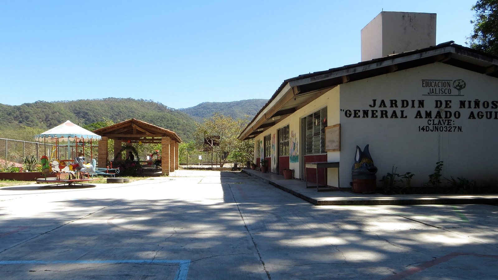 Edificio de un jardín de niños con el nombre "General Amado Aguirre" pintado en la fachada, patio y montañas boscosas al fondo