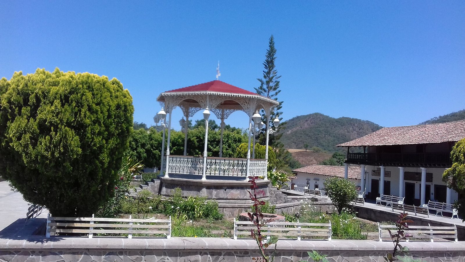 Kiosco blanco con techo rojo en la plaza de San Sebastián del Oeste, con jardines, bancas y cerros al fondo