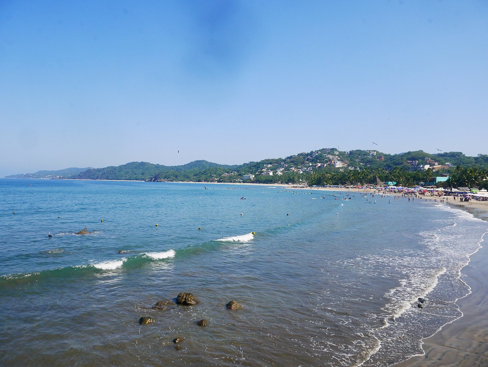 Olas pequeñas rompiendo en la bahía de Sayulita, con bañistas y surfistas en el agua y el pueblo al fondo