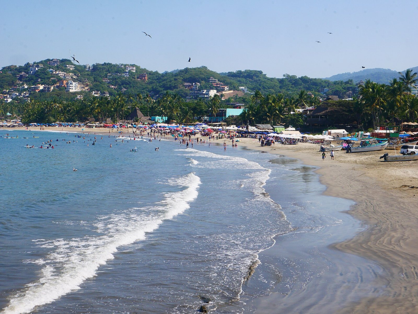 Espuma de las olas llegando a la orilla de la playa principal de Sayulita, con gente en el agua y palapas al fondo