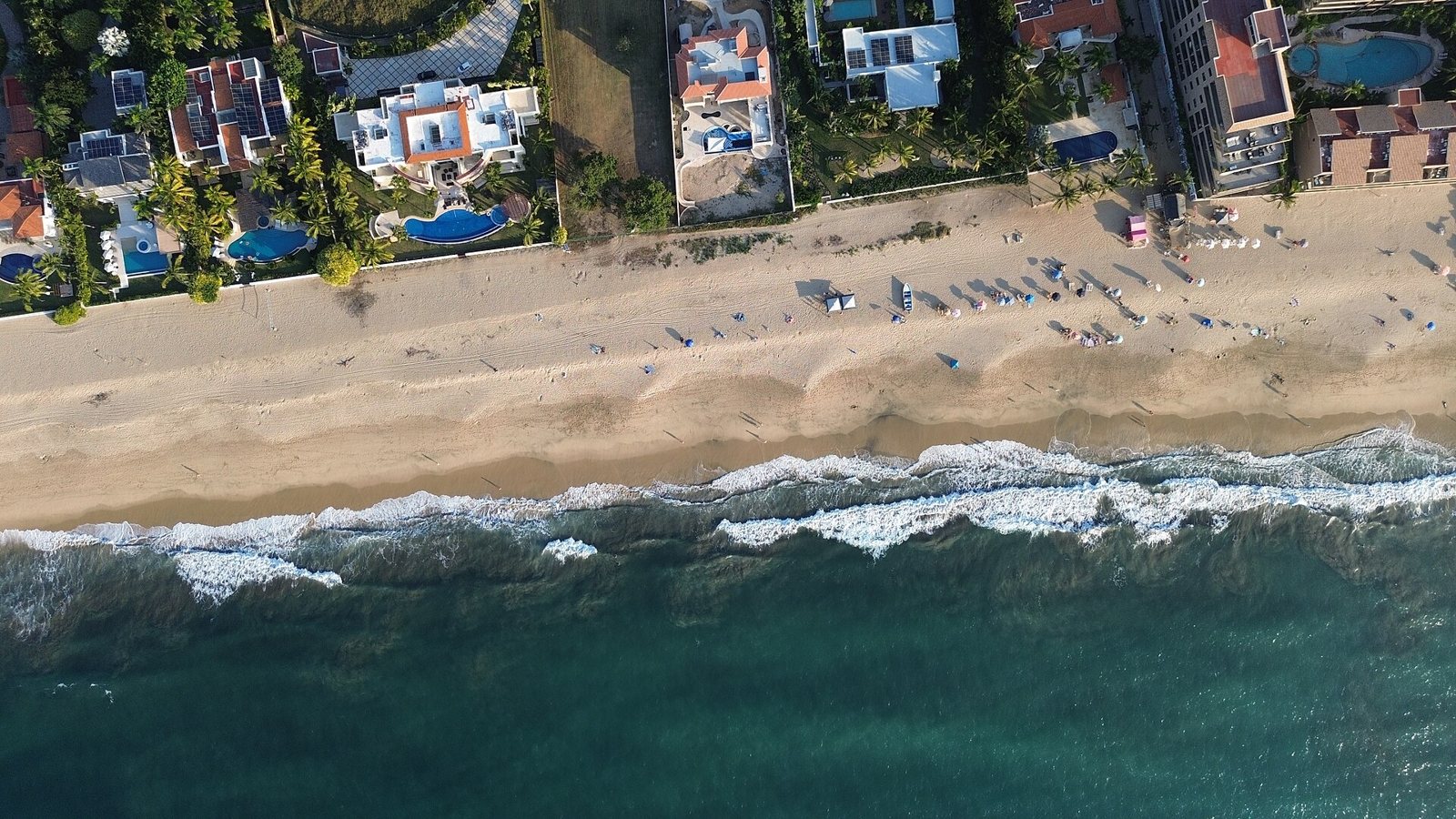 Vista aérea de las olas rompiendo sobre la arena ancha de la playa de San Pancho, con casas detrás