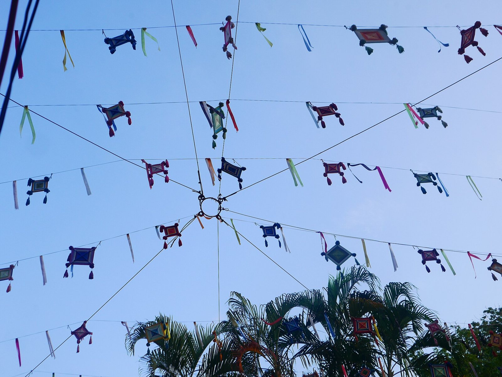 Ojos de dios tejidos de colores colgados de cuerdas sobre una calle de San Pancho, con palmeras y cielo azul