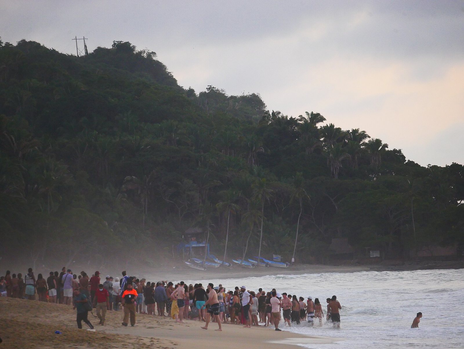 Grupo grande de personas en fila a la orilla del mar en la playa de San Pancho al atardecer, frente a un cerro con palmeras y lanchas de pescadores