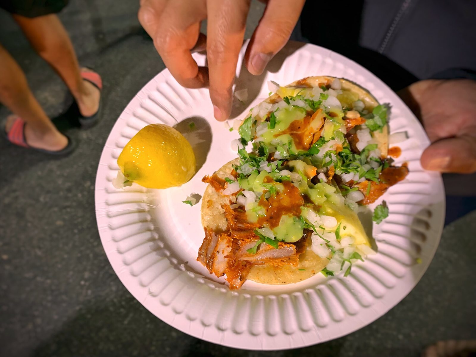 Dos tacos al pastor con cebolla, cilantro y salsas en un plato de papel, servidos de noche en la calle