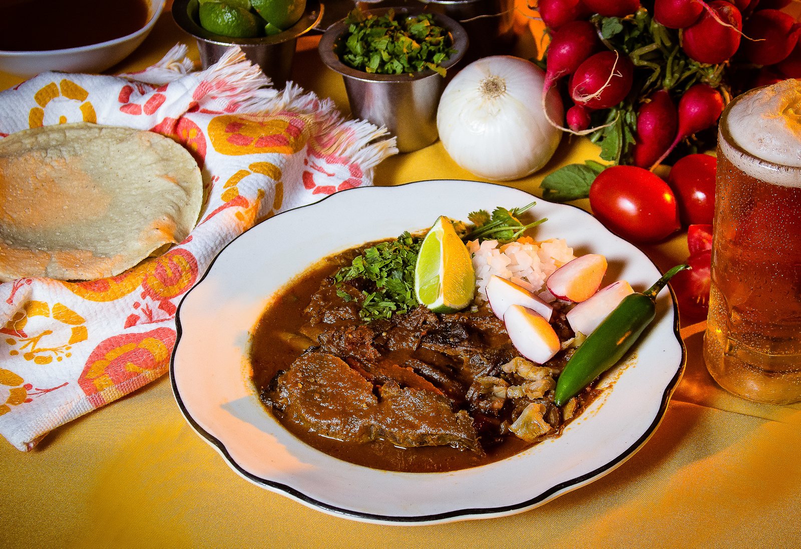 Plato de birria con cebolla, cilantro, rábano, limón y chile, junto a tortillas de maíz y un tarro de cerveza