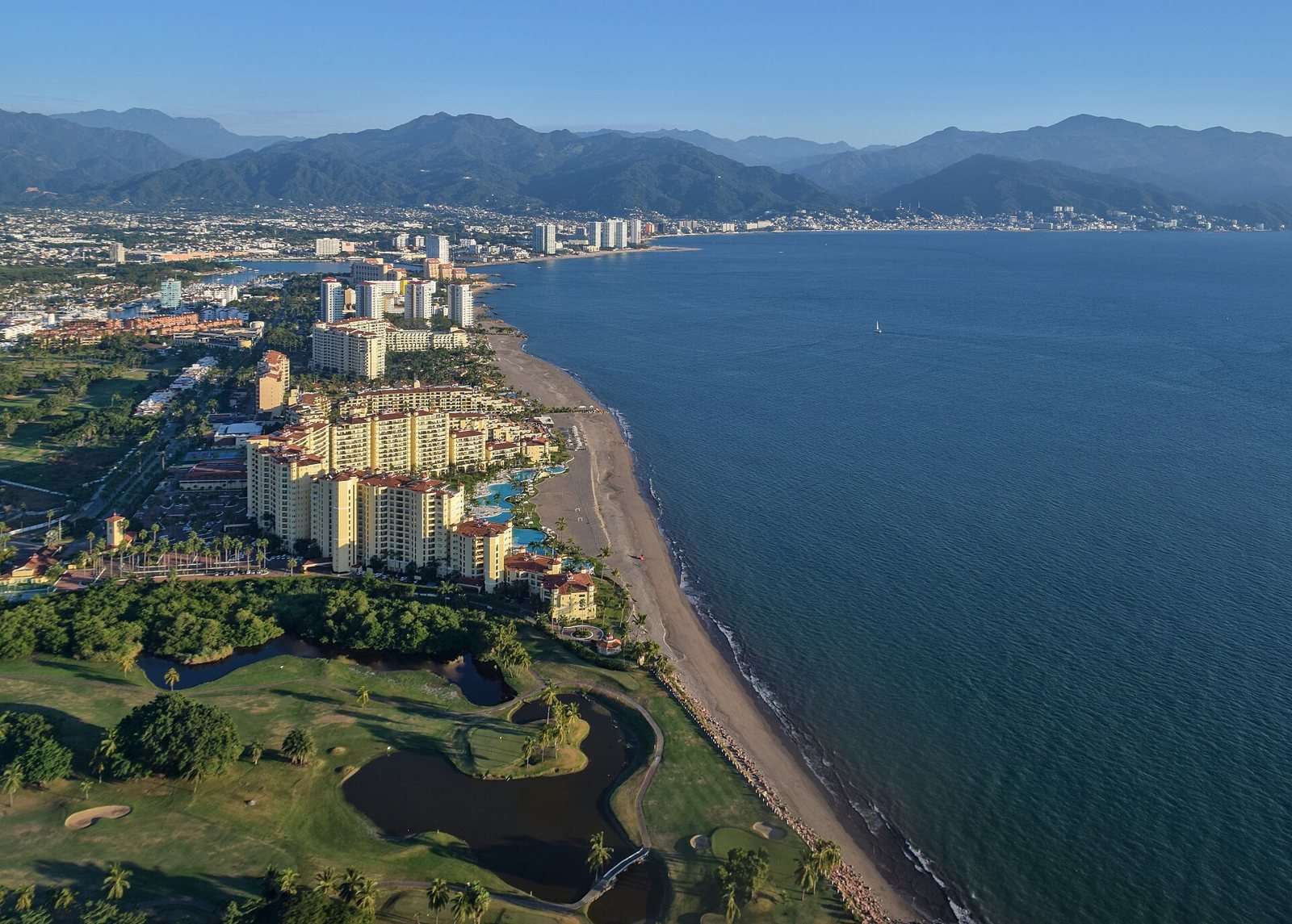 Vista aérea de la costa de Puerto Vallarta con torres de hoteles junto a la playa, un campo de golf en primer plano y la sierra al fondo