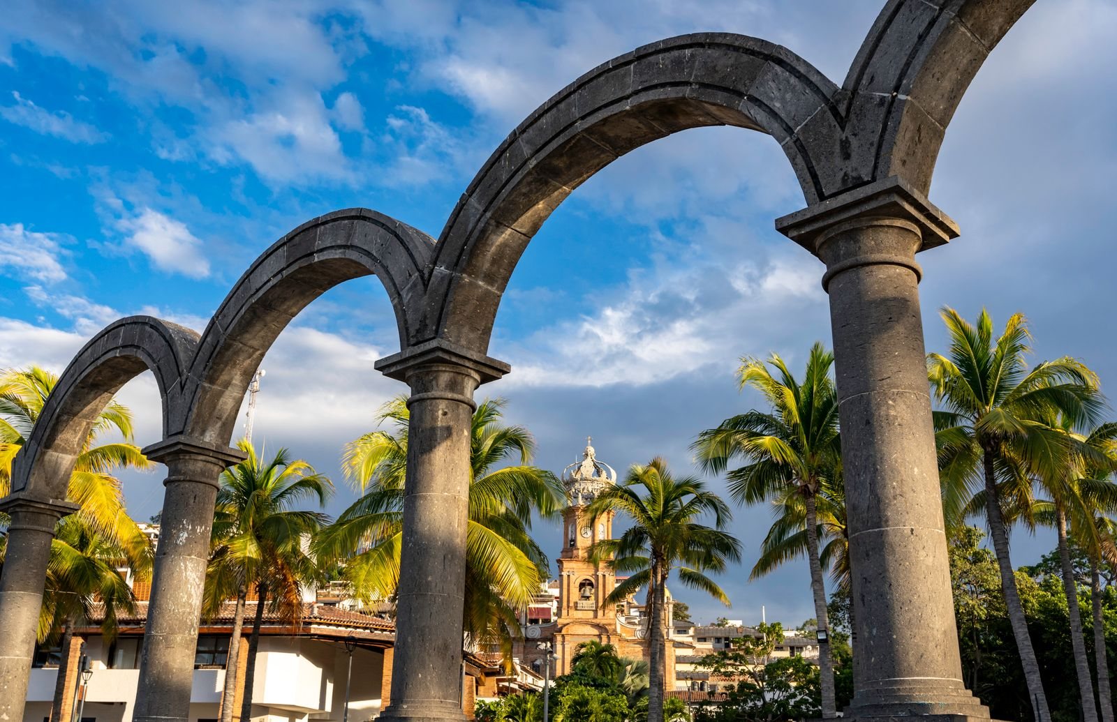 Arcos de piedra del Malecón de Puerto Vallarta con palmeras y la torre con corona de la parroquia de Guadalupe al fondo