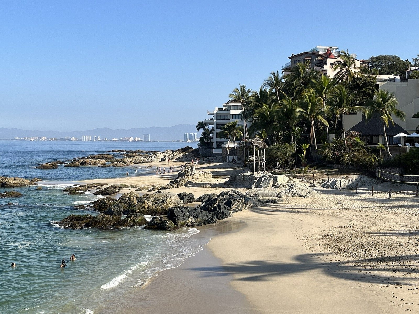 Playa de arena clara con rocas, palmeras y edificios en la ladera, con la zona hotelera de Vallarta a lo lejos