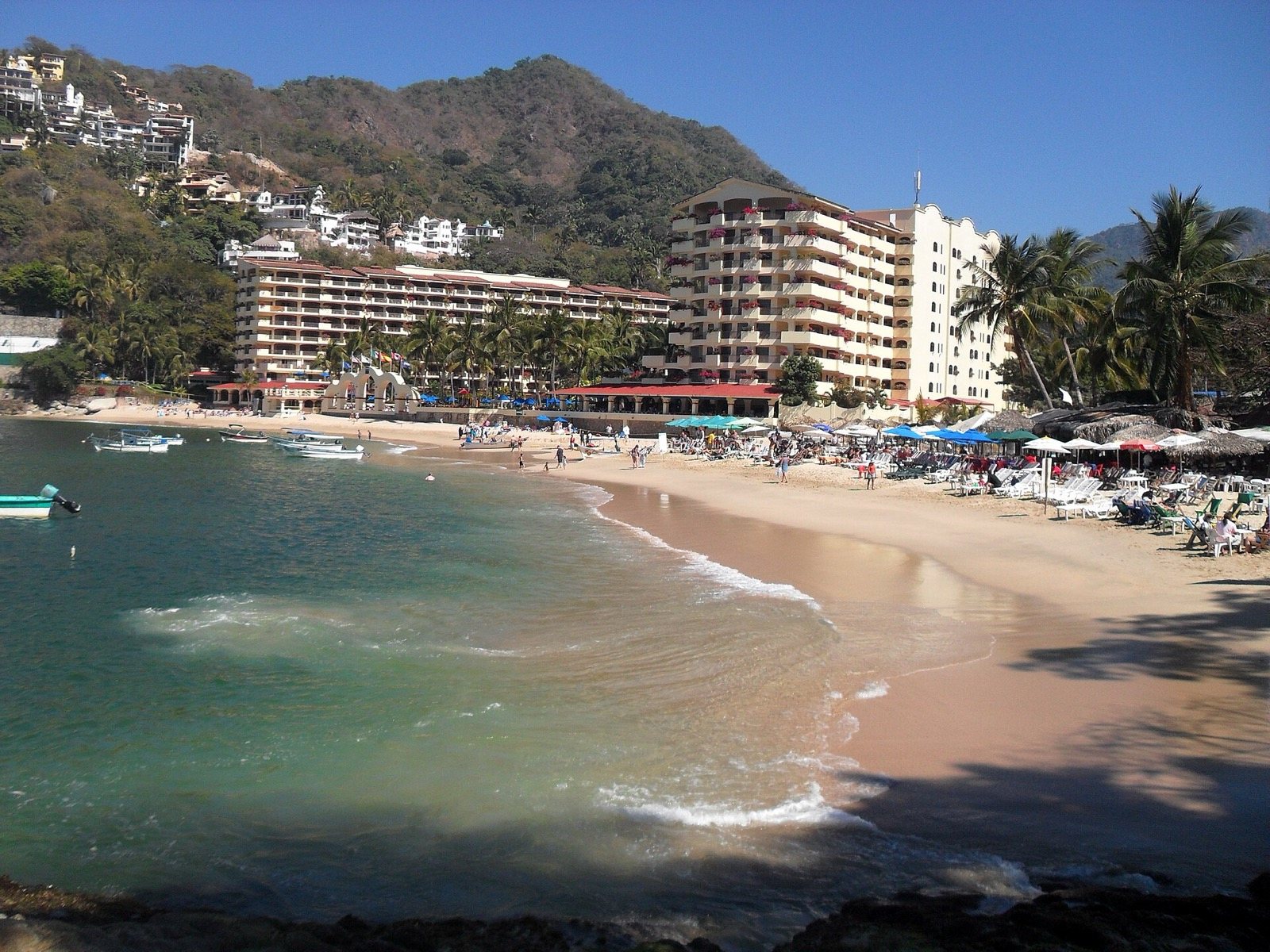 Playa de Mismaloya en forma de media luna con lanchas, palapas, edificios de hotel frente al mar y cerros verdes al fondo