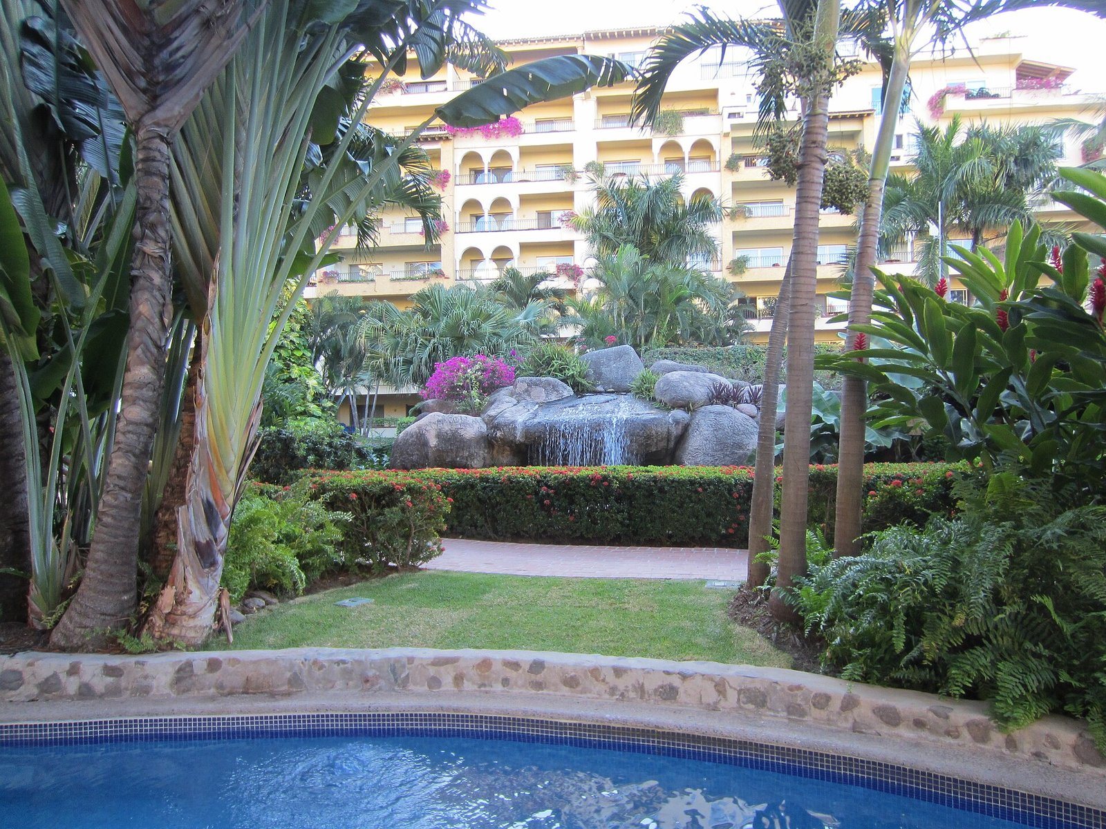 Jardín tropical con palmeras, rocas y una cascada junto a una alberca, con el edificio del hotel al fondo