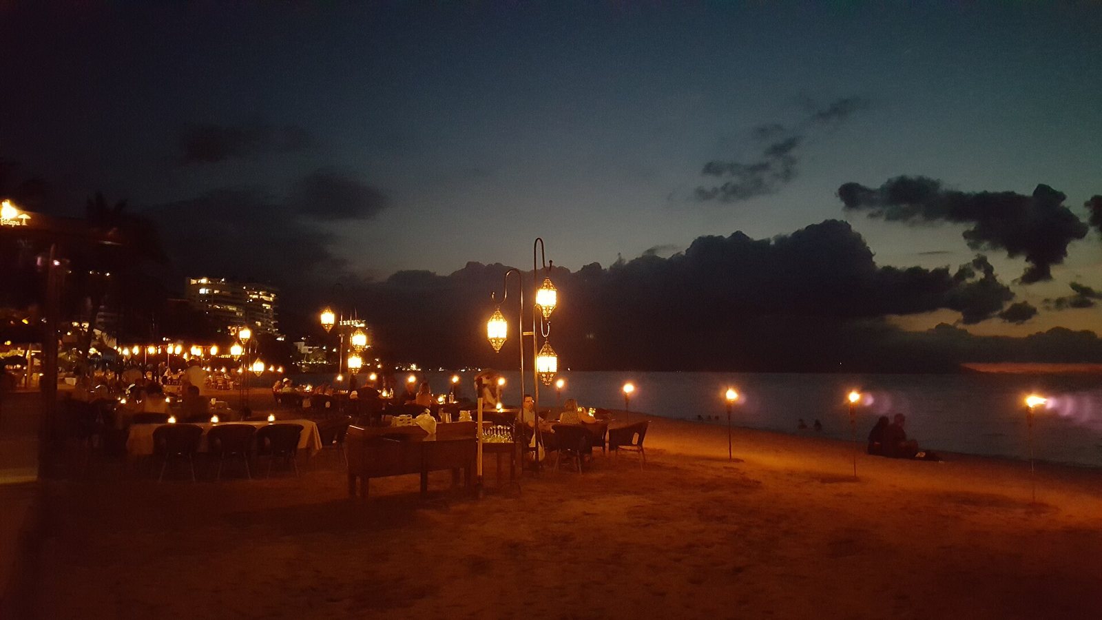 Mesas iluminadas con faroles y antorchas sobre la arena frente al mar, ya de noche, en Playa Los Muertos
