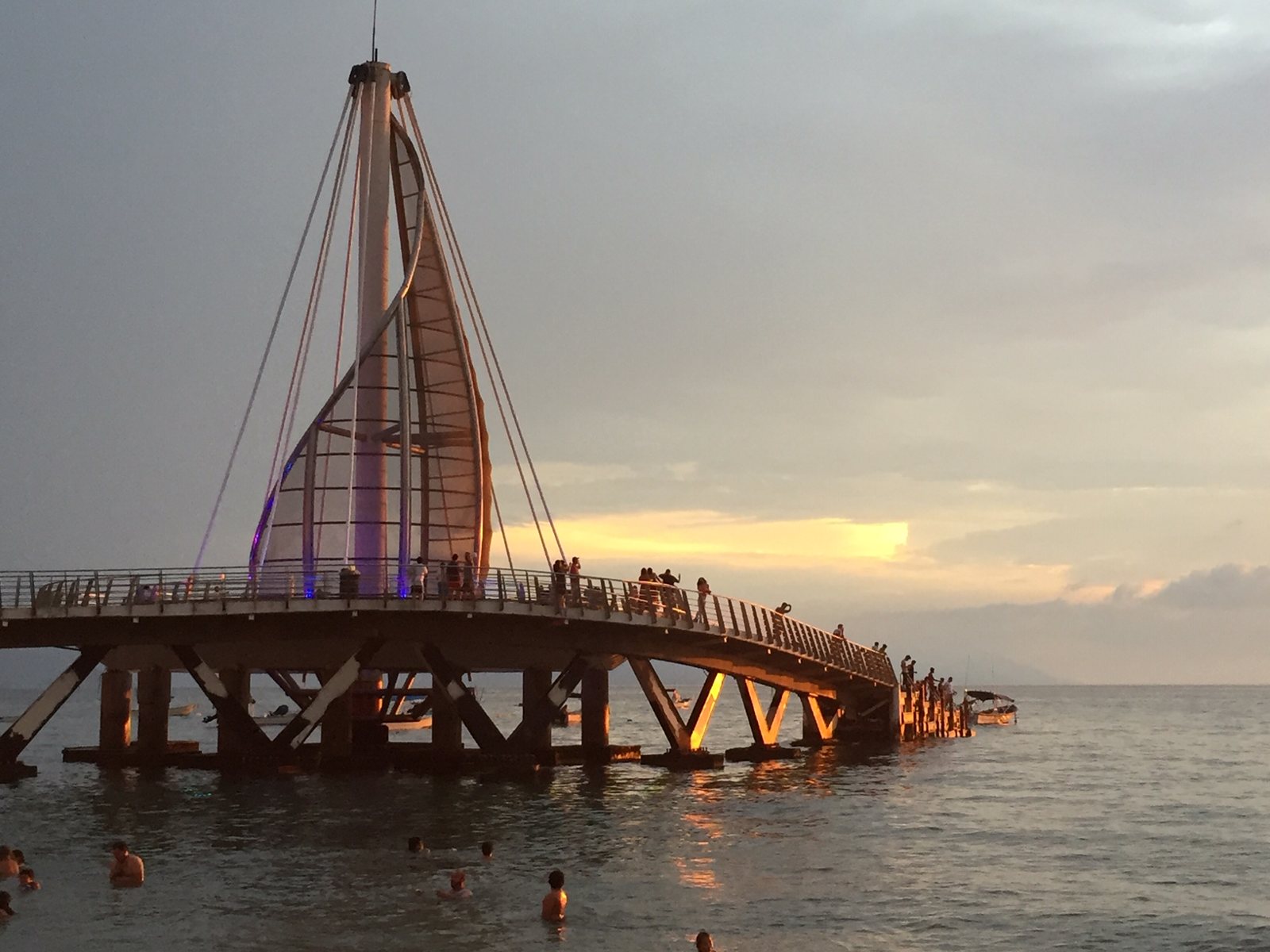Muelle de Playa Los Muertos con su estructura en forma de vela al atardecer, con personas caminando sobre él y nadando en el mar