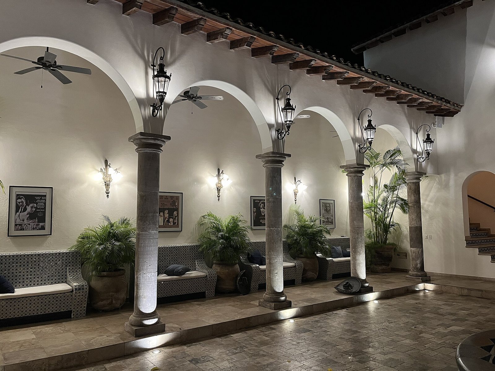 Corredor de arcos iluminado de noche con columnas de cantera, faroles, bancas de azulejo, palmas en macetas y carteles de películas de Elizabeth Taylor y Richard Burton en Casa Kimberly