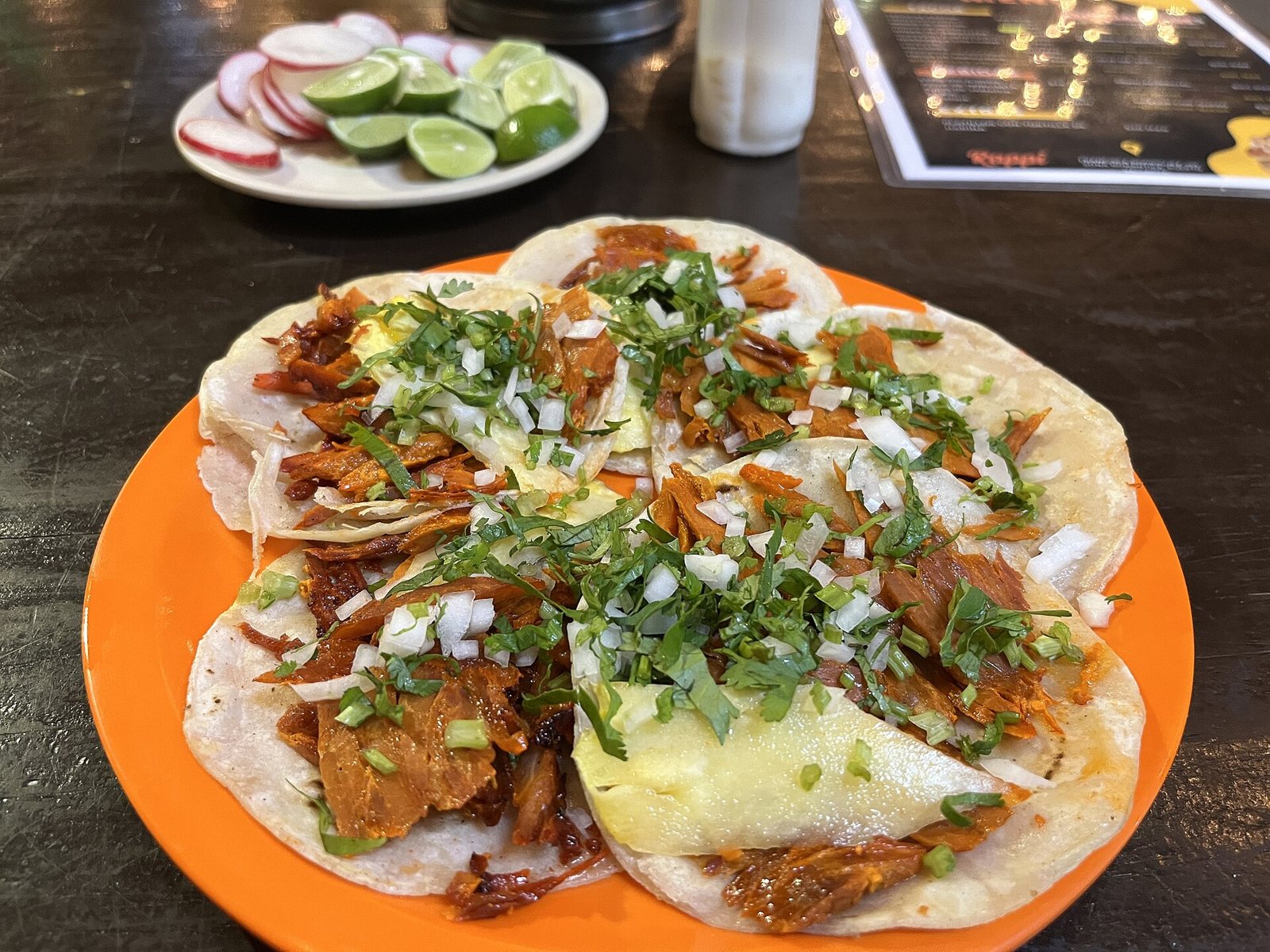 Plato naranja con seis tacos al pastor con piña, cebolla y cilantro, y al fondo un plato con limones y rábanos