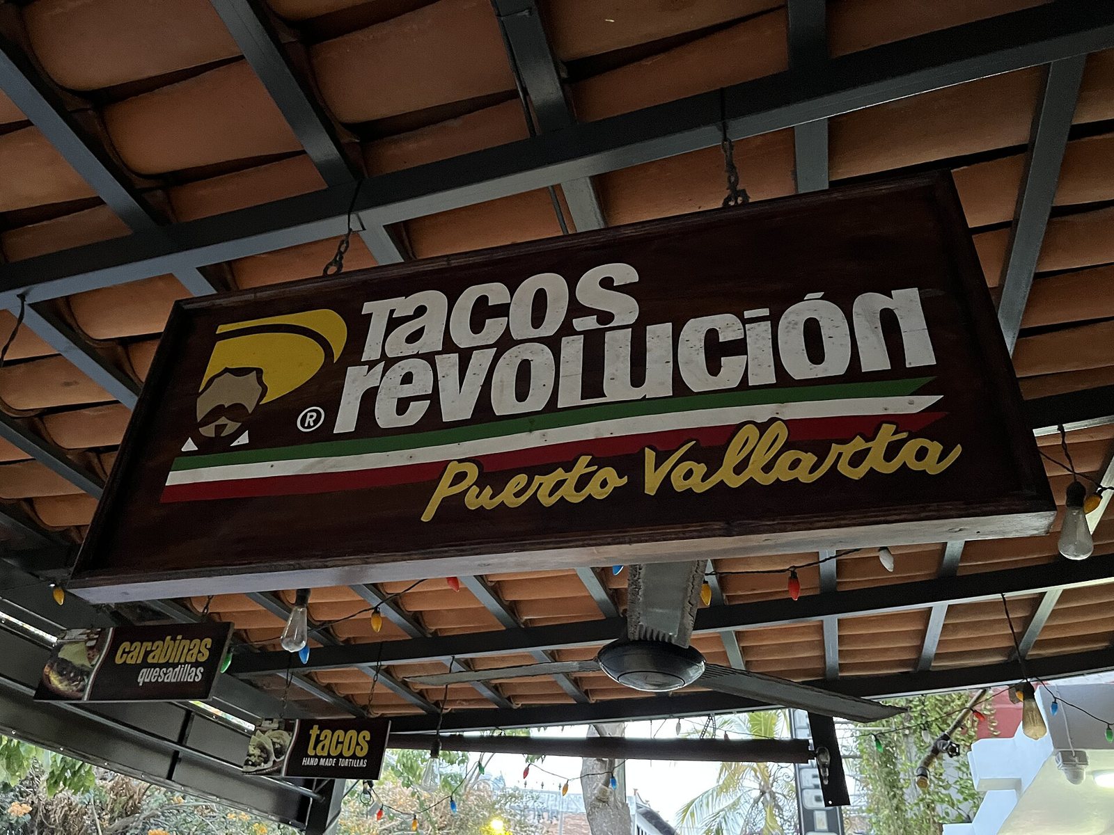 Letrero de madera de Tacos Revolución Puerto Vallarta con franja tricolor, colgado bajo un techo de teja con series de luces