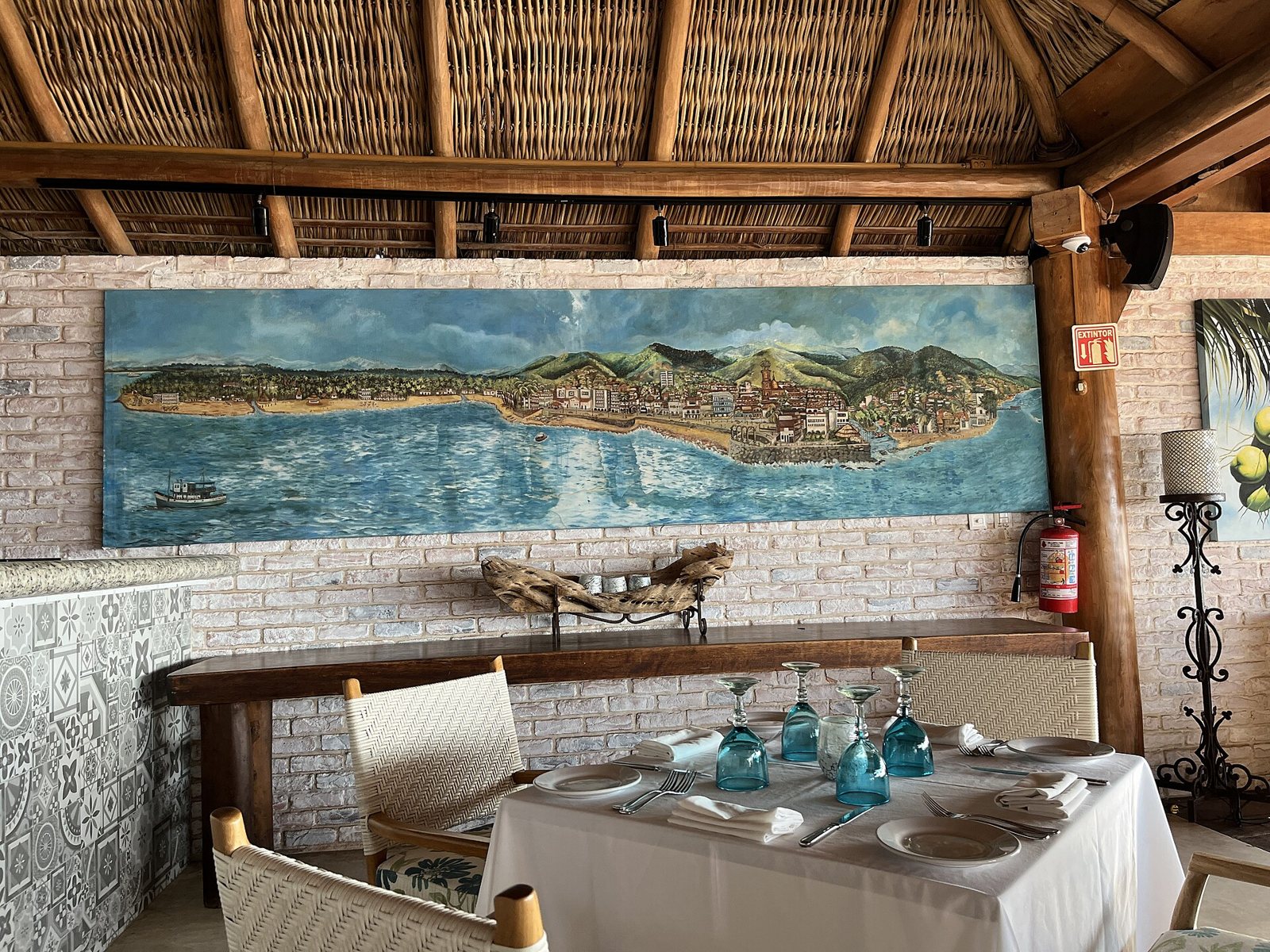Mesa con mantel blanco y copas azules bajo un techo de palapa, con un mural de la bahía de Puerto Vallarta en la pared