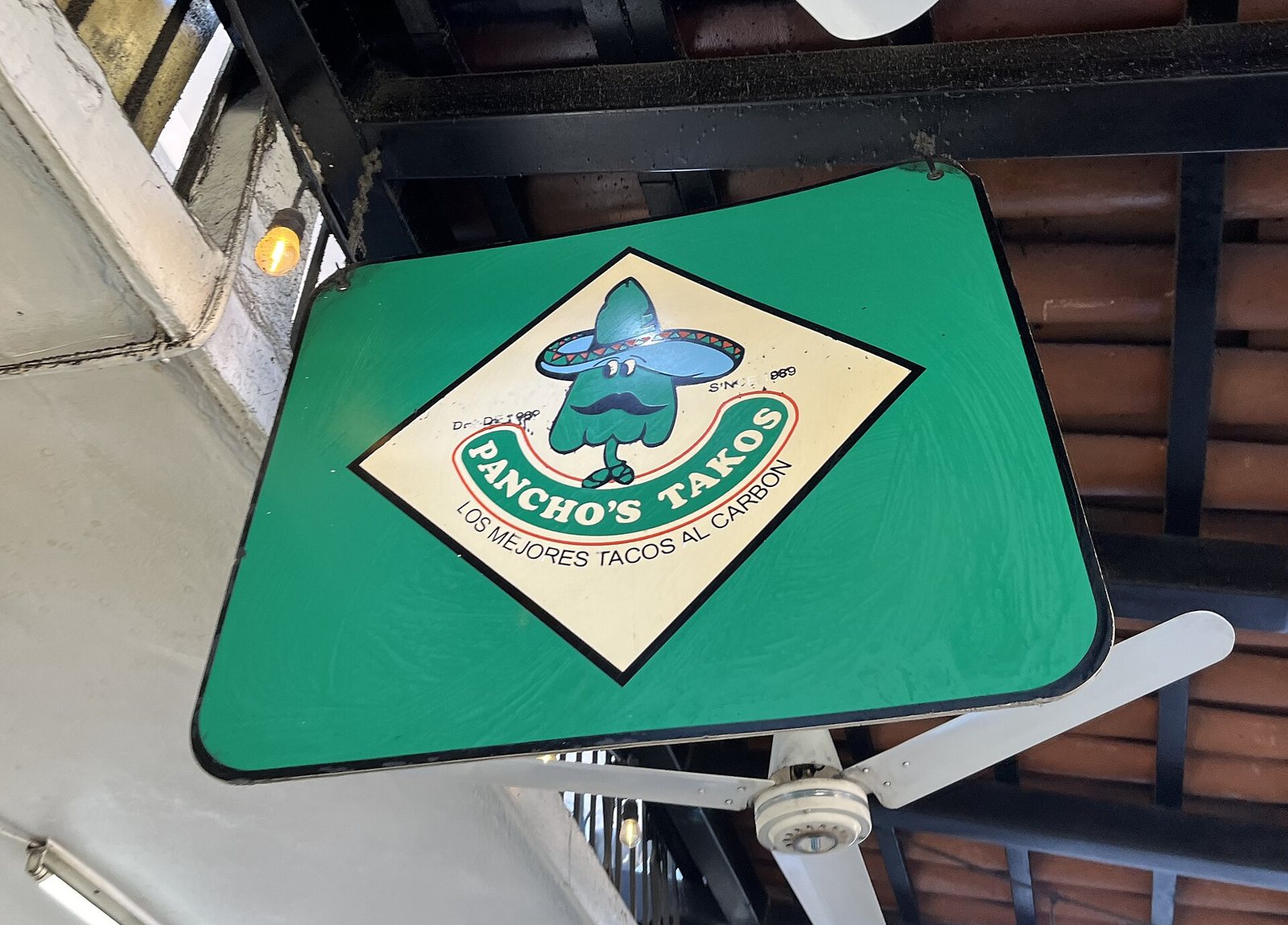 Letrero verde colgante de Pancho's Takos con la leyenda "Los mejores tacos al carbón", bajo un techo de teja