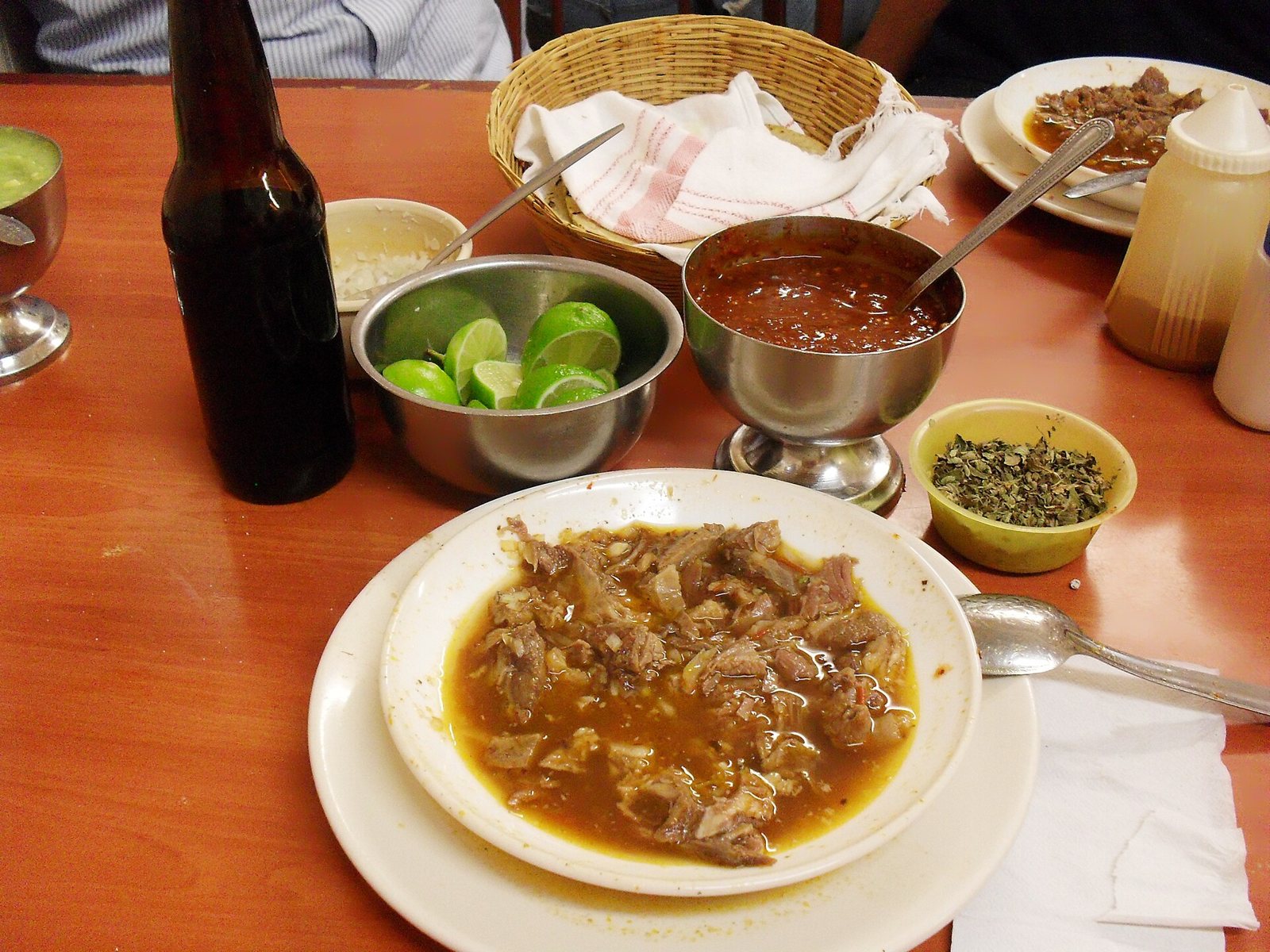 Plato de birria sobre una mesa con limones, salsa roja, orégano, cebolla y una canasta de tortillas