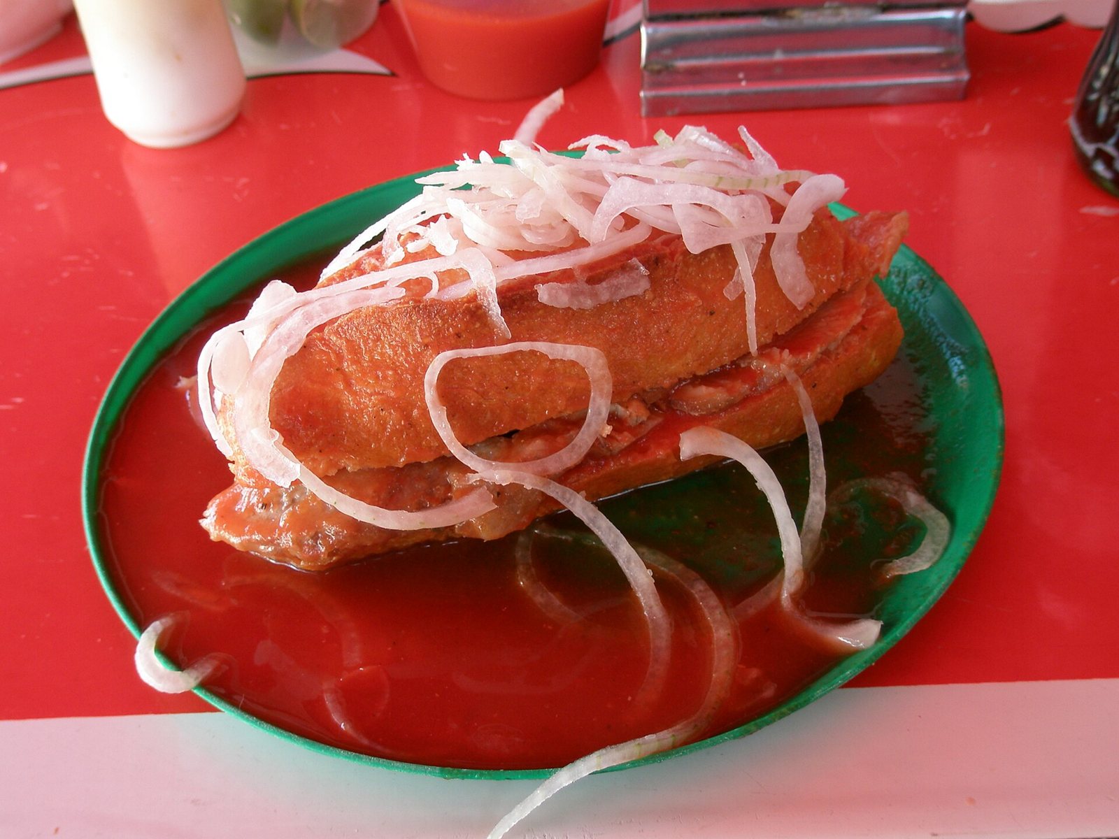 Torta ahogada bañada en salsa roja de jitomate en un plato verde, con cebolla en rodajas encima