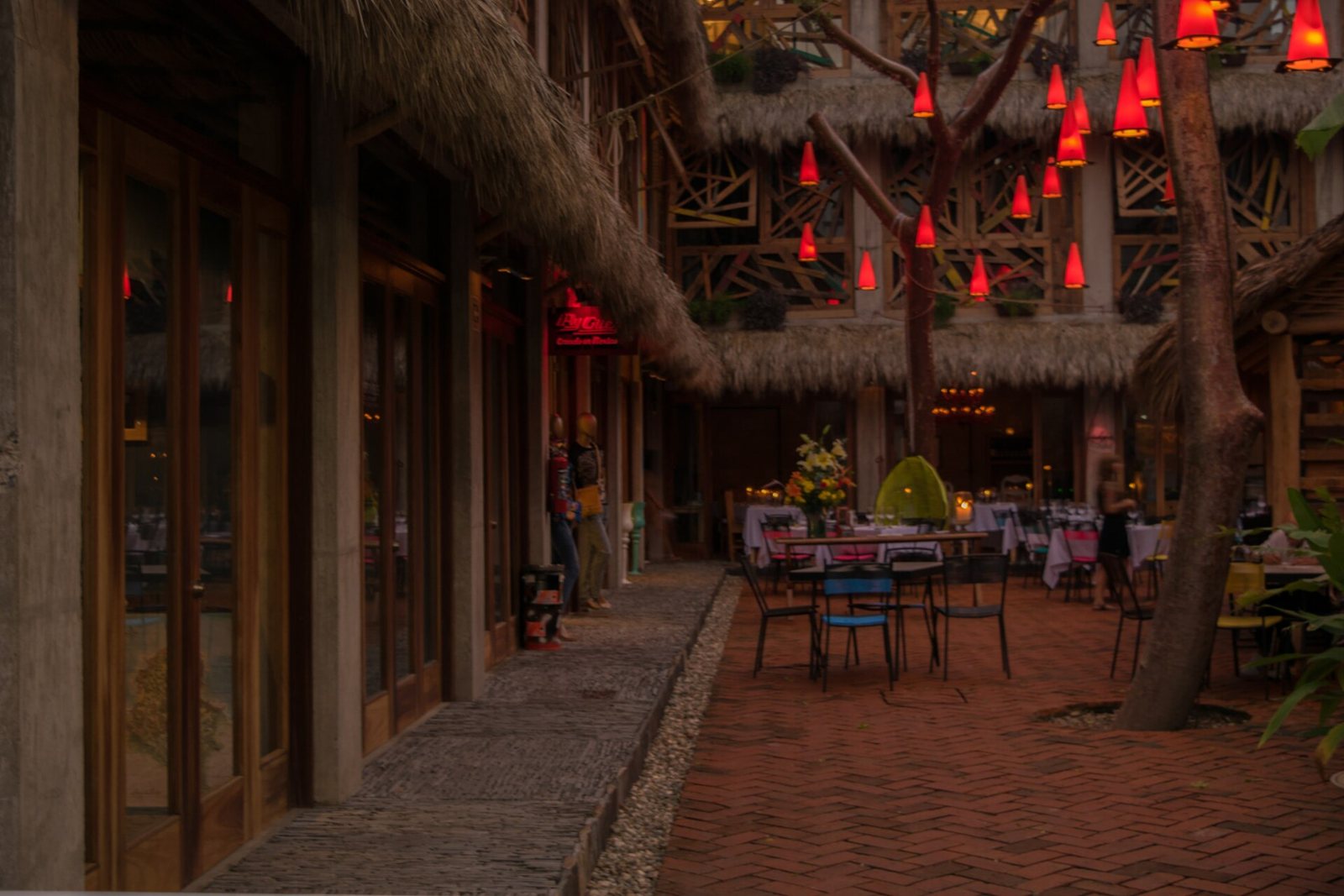 Patio de un restaurante en Sayulita al anochecer, con mesas al aire libre, techos de palapa, piso de ladrillo y lámparas rojas colgando de un árbol