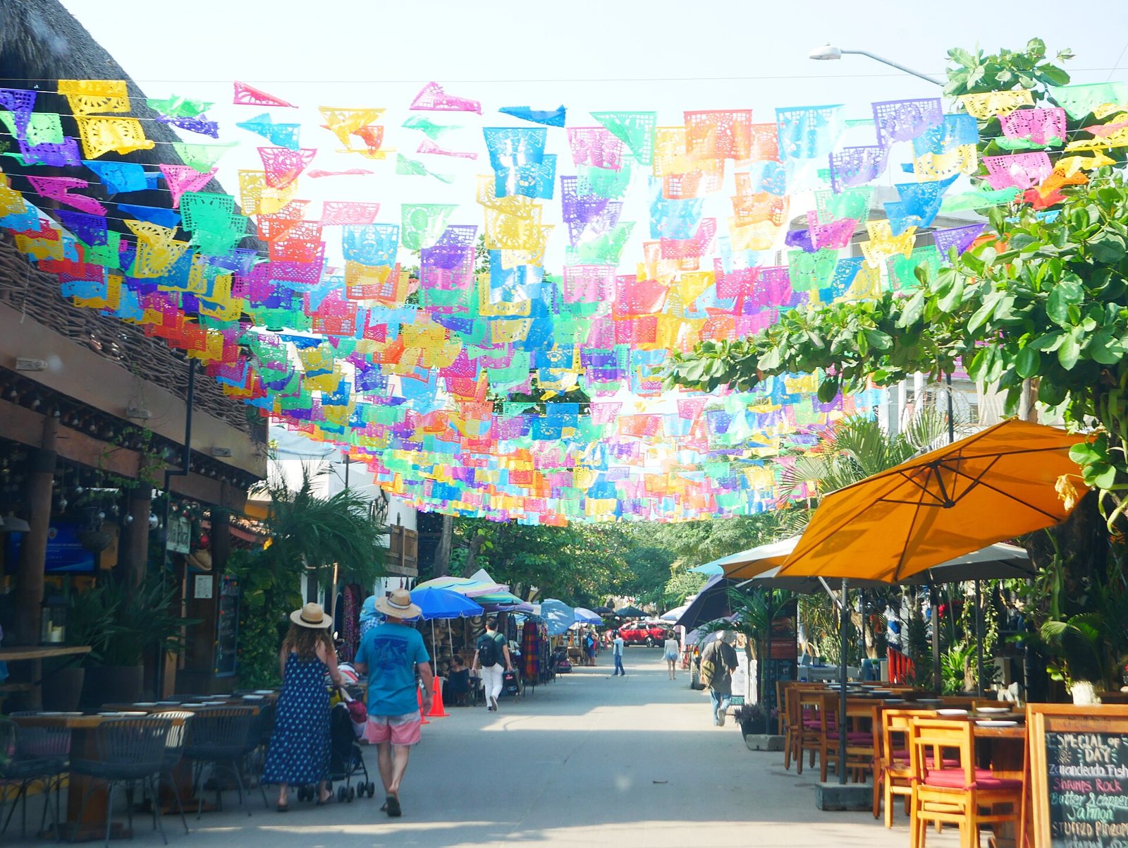 Avenida Tercer Mundo de San Pancho cubierta de papel picado, con mesas y sombrillas de restaurantes a un lado de la calle y gente caminando