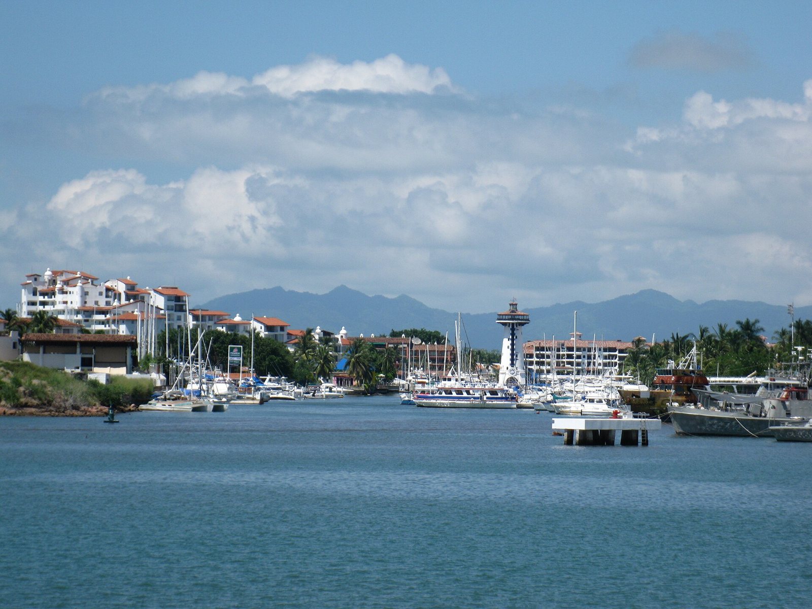 Vista de Marina Vallarta desde el agua: veleros y yates, edificios blancos, la torre del faro al centro y la Sierra Madre al fondo